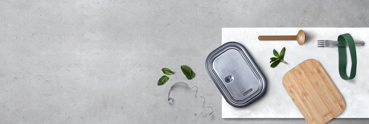 Replacement lids and spoons for reusable lunch boxes on a grey bench with marble chopping board